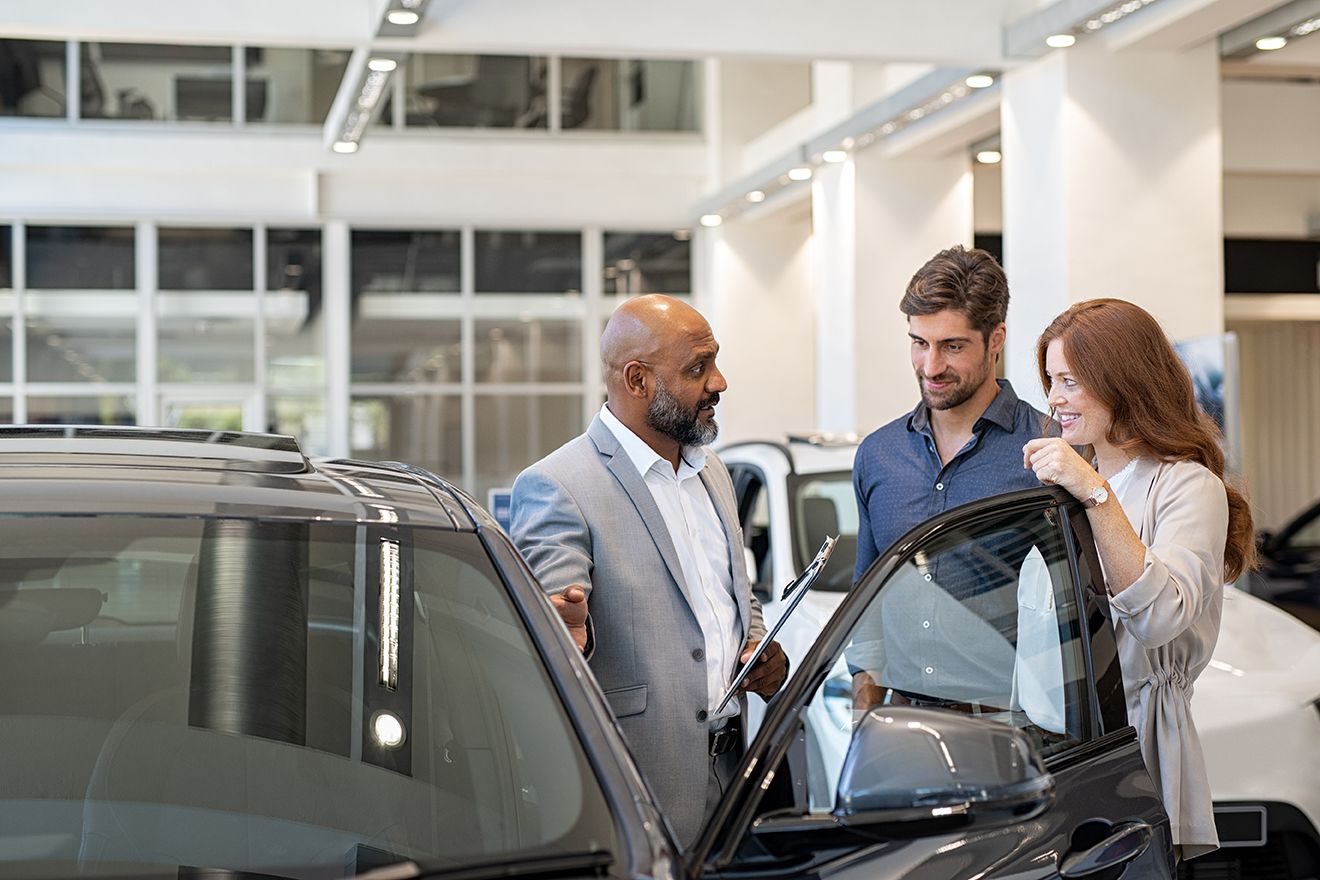 Beautiful couple in casual clothes smiling while examining a new car in a motor show with salesman. Mature salesman showing new luxury car to man and woman in showroom. Happy couple with cardealer showing new features in auto dealership.